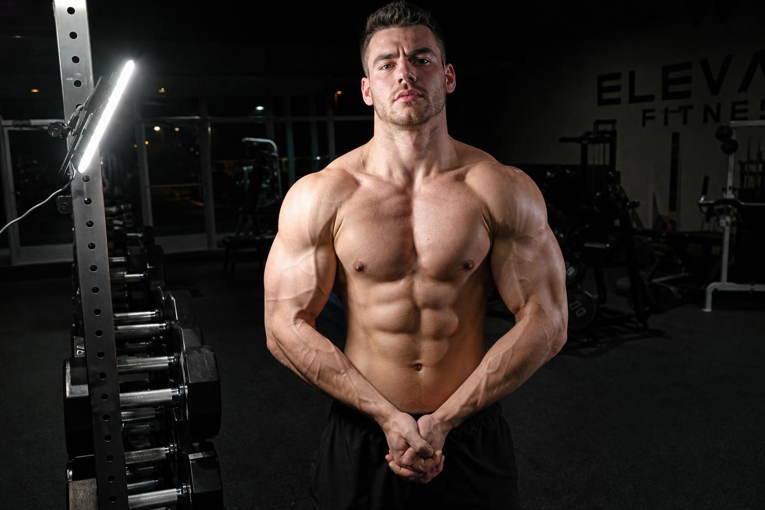 The same man flexing in the same gym, lit at 45 degrees by a GYMLIGHT bar clamped to the dumbbell rack upright, showing vascular arms, sharp pec-to-shoulder separation and real shadow under the abdominals
